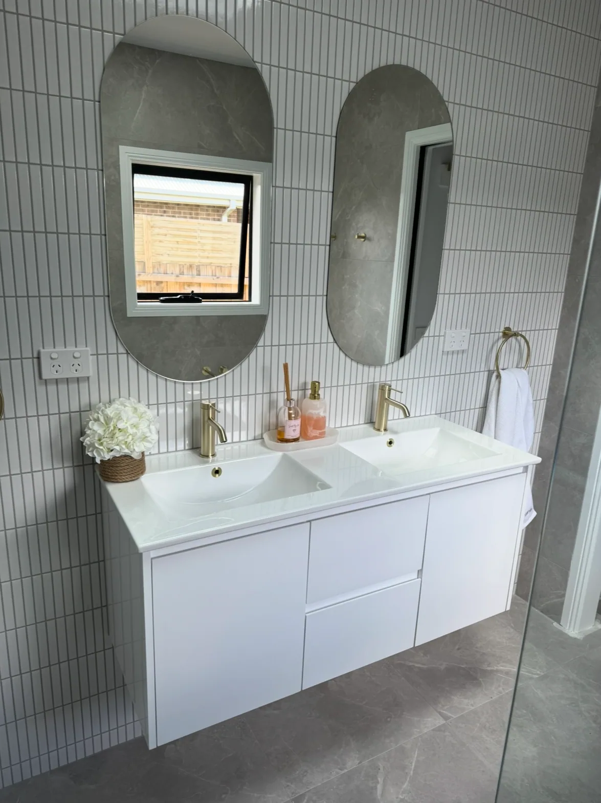 Floating white double vanity with two brass basin mixers and oval mirrors on a white vertical finger-tile wall