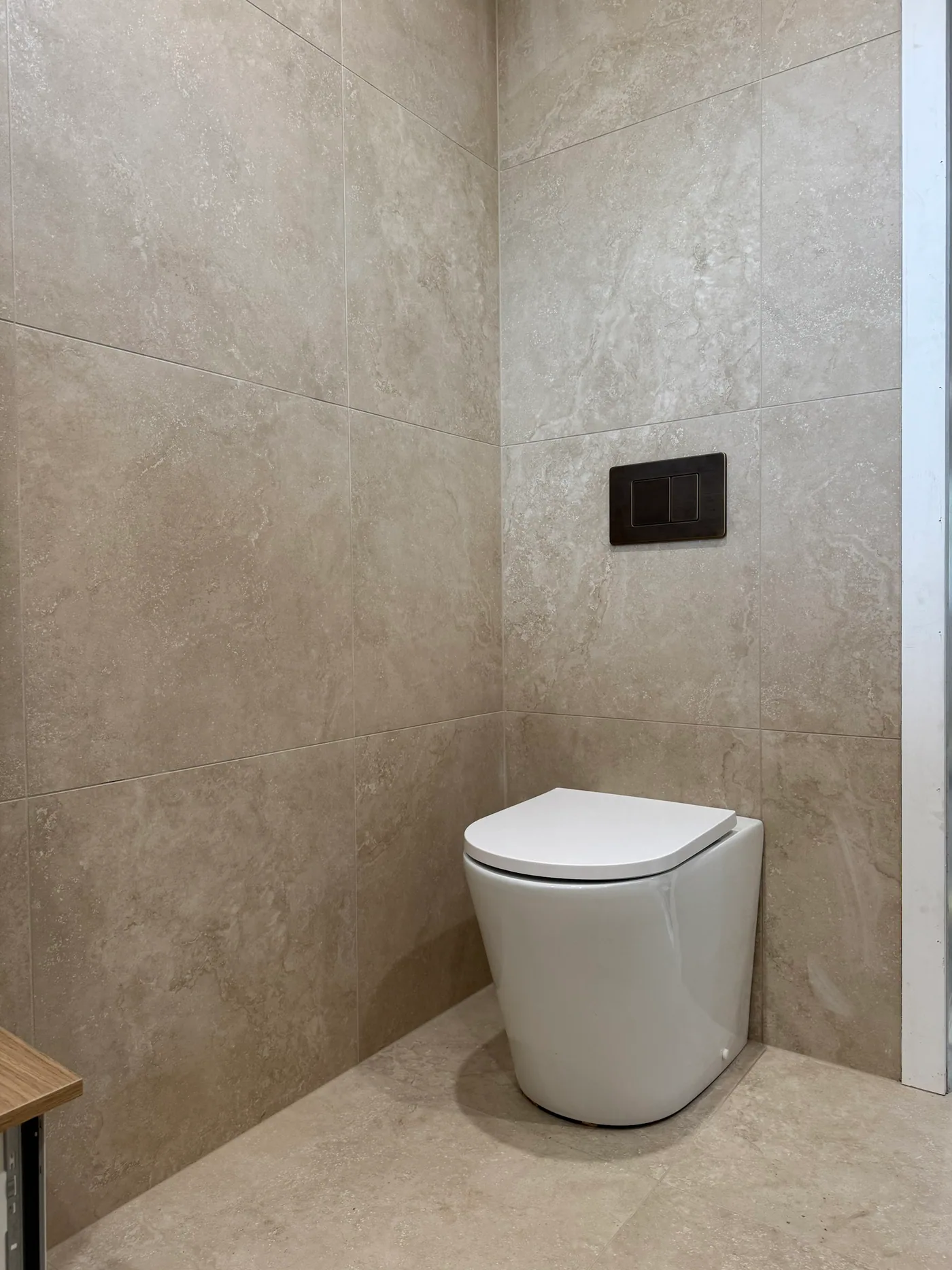 White wall-faced toilet pan with a matte black dual flush plate set into travertine-look wall tiles